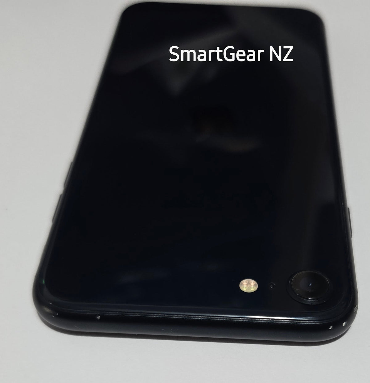 A black smartphone lying on a white surface, viewed from above. The back of the phone is glossy black with a small camera lens and flash visible near the bottom left.