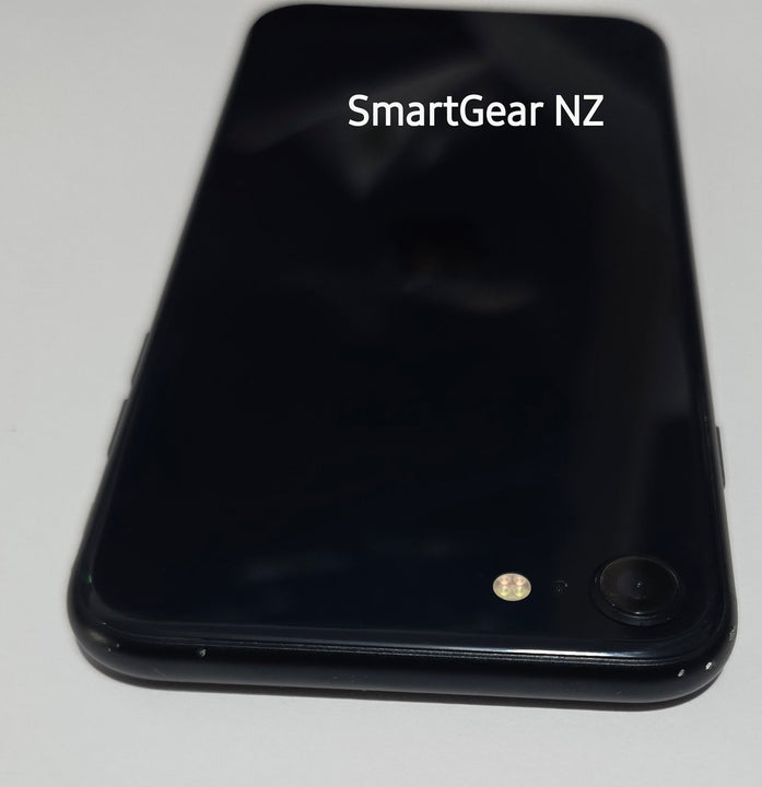 A black smartphone lying on a white surface, viewed from above. The back of the phone is glossy black with a small camera lens and flash visible near the bottom left.