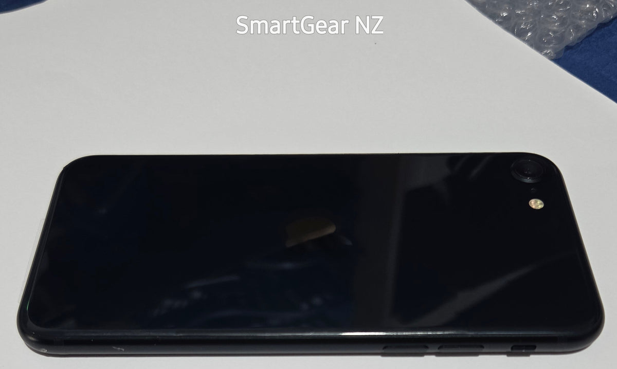 A black smartphone shown from the back, resting on a white surface with blue fabric around it. The text 'SmartGear NZ' is visible near the top.