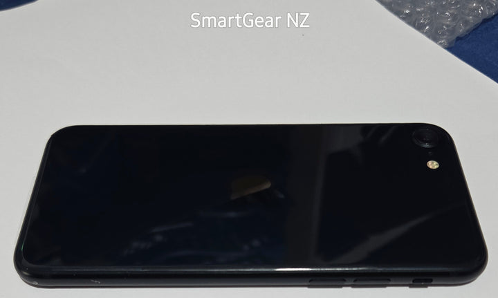 A black smartphone shown from the back, resting on a white surface with blue fabric around it. The text 'SmartGear NZ' is visible near the top.