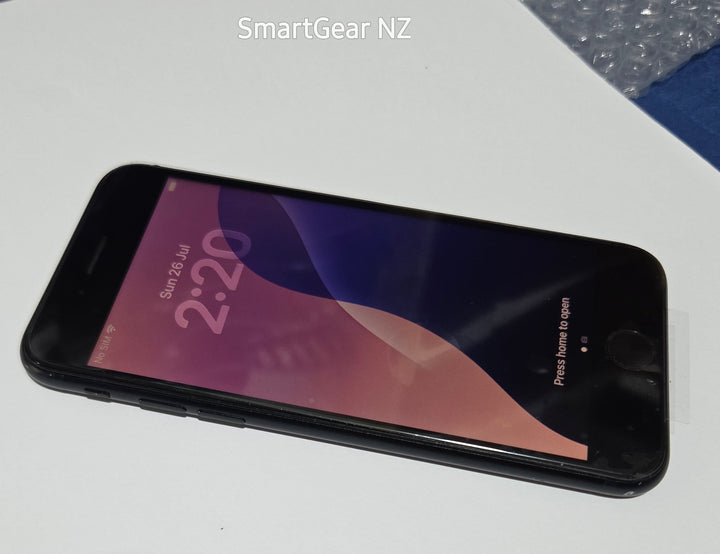 A black smartphone lying on a white surface, with a blue textured fabric and bubble wrap visible in the background. The screen is on and displays a pink and purple wallpaper with the time 2:20.