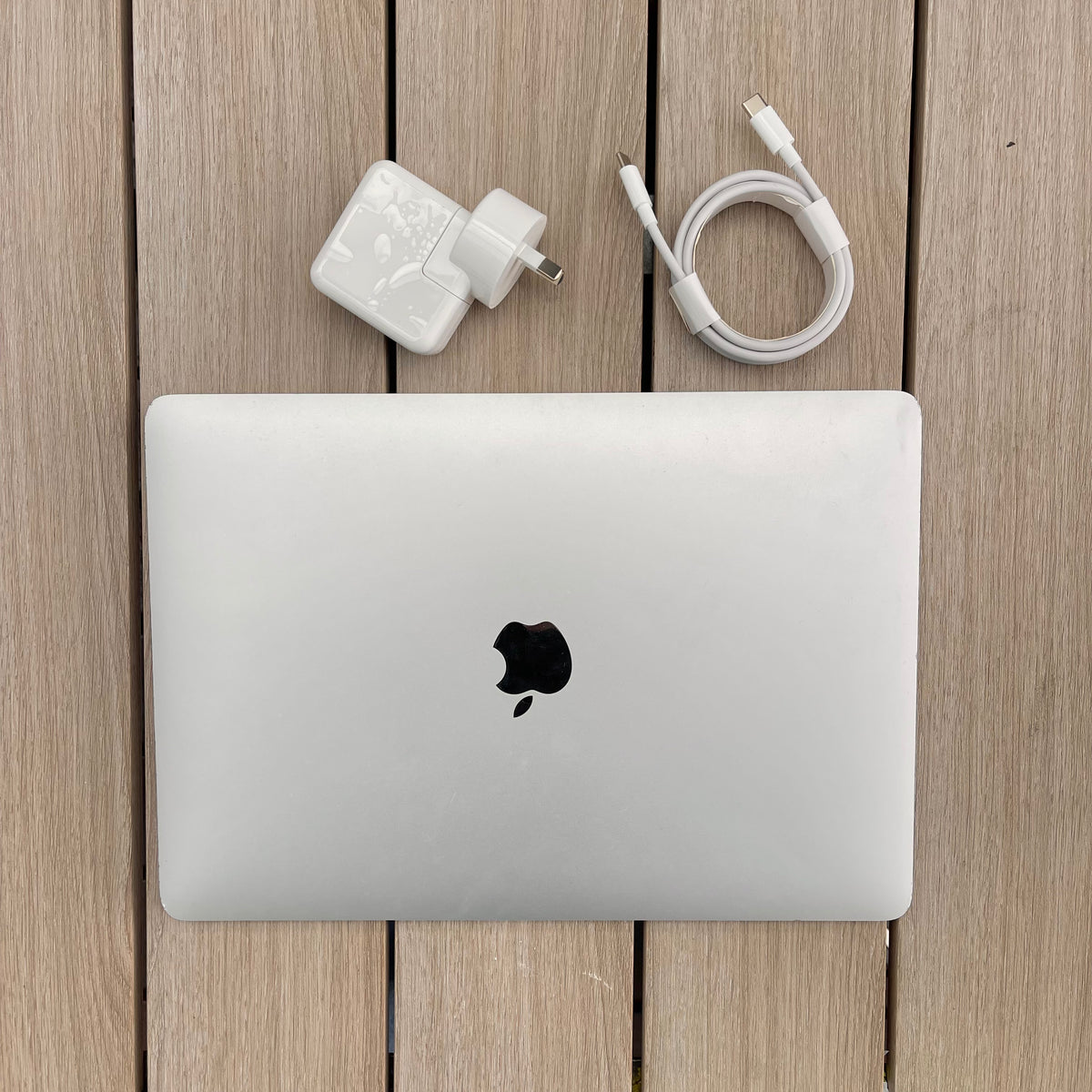 MacBook Air (Retina, 13-inch, 2018) in silver color with a few marks and dents, displayed alongside a white 30W USB-C Power Adapter and a USB-C Charge Cable, all placed on a wooden surface.